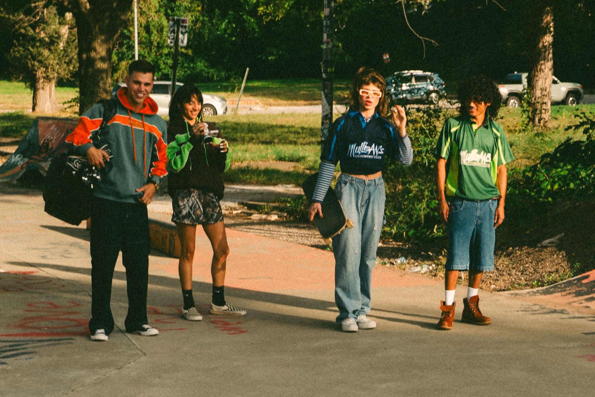 Four friends at a skatepark in the sun: two in Mulligan's Country Club jerseys, one holding a skateboard, one in an orange and teal hoodie